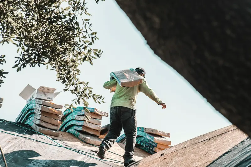 A roofer carrying a bundle of shingles across a residential roof